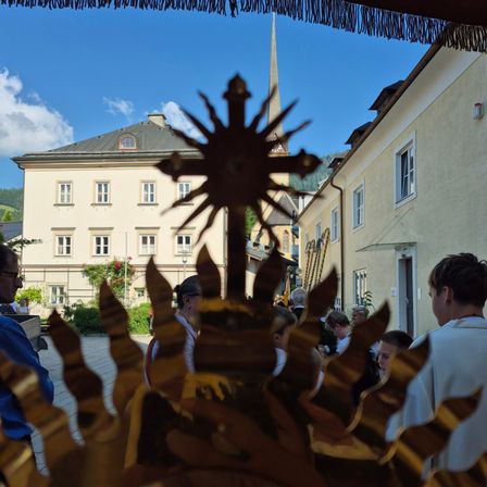 Eine Gruppe von Menschen versammelt sich um ein goldenes Sonnendesign, mit einer Kirche im Hintergrund.