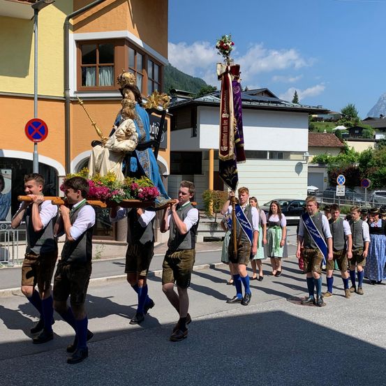 Eine Gruppe von Menschen in traditioneller Kleidung trägt einen geschmückten Festwagen mit einer Statue und Blumen. Sie gehen in einer Prozession auf einer Straße, mit Gebäuden und Bergen im Hintergrund.