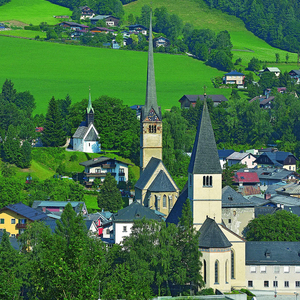 Ein Dorf in einem grünen Tal mit einer Kirche mit einem hohen Turm und kleineren Spitzen. Das Dorf hat viele Häuser und Bäume.