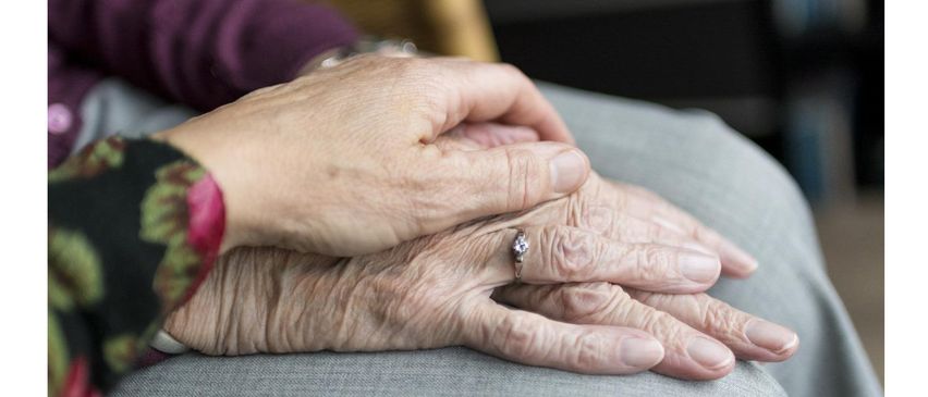 Eine ältere Person mit einem Ring am Finger hält die Hand einer anderen Person, möglicherweise einer Pflegekraft, die ebenfalls ihre Hände hält. Beide sitzen auf einem Stuhl, wobei im Hintergrund ein Bücherregal und ein Holzstuhl zu sehen sind. Das Bild ist Teil einer Veranstaltungsankündigung für Vorträge von Dr. Simone Pölzer über Demenz und Delirium.