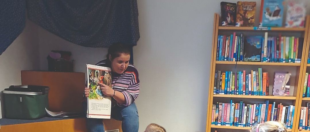 Eine Frau liest ein Buch vor einer Gruppe von Kindern, die im Kreis auf dem Bibliotheksboden sitzen. Die Kinder lauschen aufmerksam. Im Hintergrund befinden sich mit Büchern gefüllte Regale.