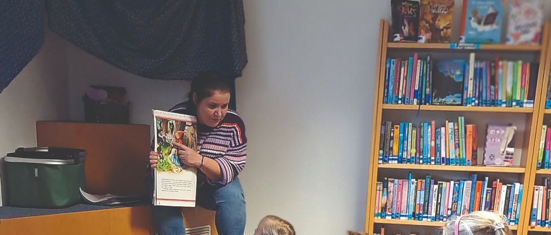 Eine Frau liest ein Buch vor einer Gruppe von Kindern vor, die im Lesesaal auf dem Boden sitzen. Die Kinder hören aufmerksam zu. Dahinter befinden sich mit Büchern gefüllte Regale.