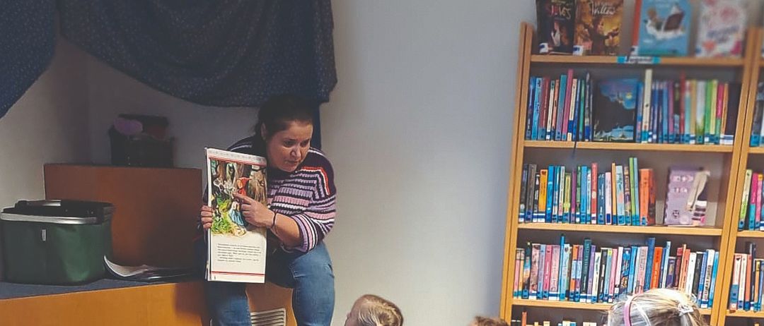 Eine Frau liest ein Buch für eine Gruppe von Kindern vor, die auf dem Boden in einer Bibliothek sitzen. Die Kinder lauschen aufmerksam der Geschichte.