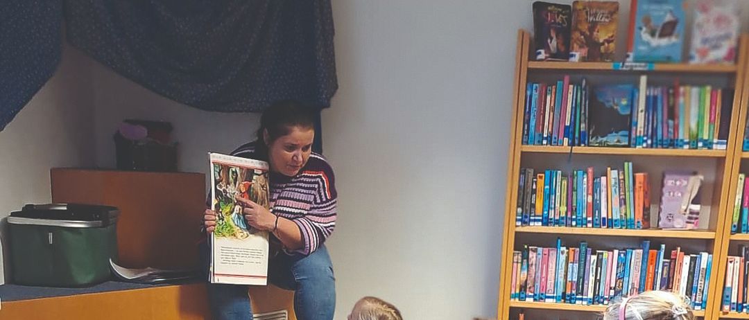 Eine Frau liest Kindern in einer Bibliothek ein Buch vor. Einige sitzen auf Kissen, andere auf dem Boden. Bücherregale und Aufbewahrungsboxen sind im Hintergrund.