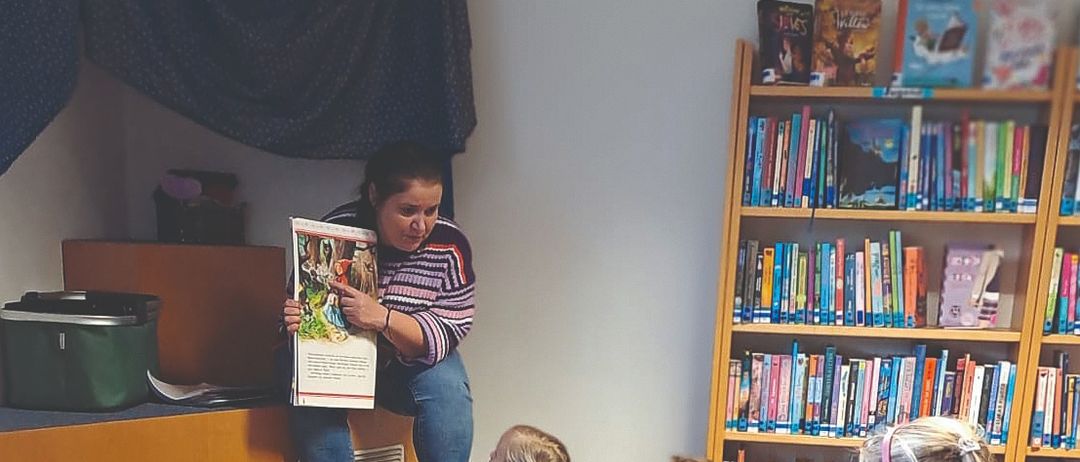 Eine Frau liest ein Buch vor einer Gruppe von Kindern, die in einer Bibliothek auf dem Boden sitzen.