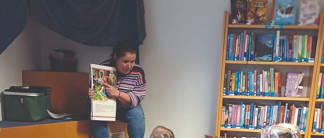 Eine Frau liest ein Buch für eine Gruppe von Kindern vor, die in einem Kreis auf dem Bibliotheksboden sitzen, umgeben von bunten Teppichen und Bücherregalen.
