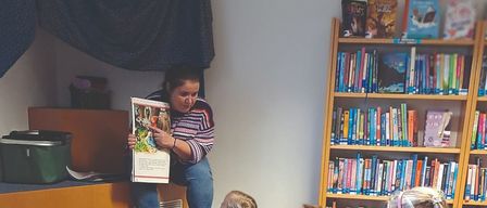 A woman reads a book to a group of children sitting on the floor in a library. Colorful rugs and shelves filled with books are visible.