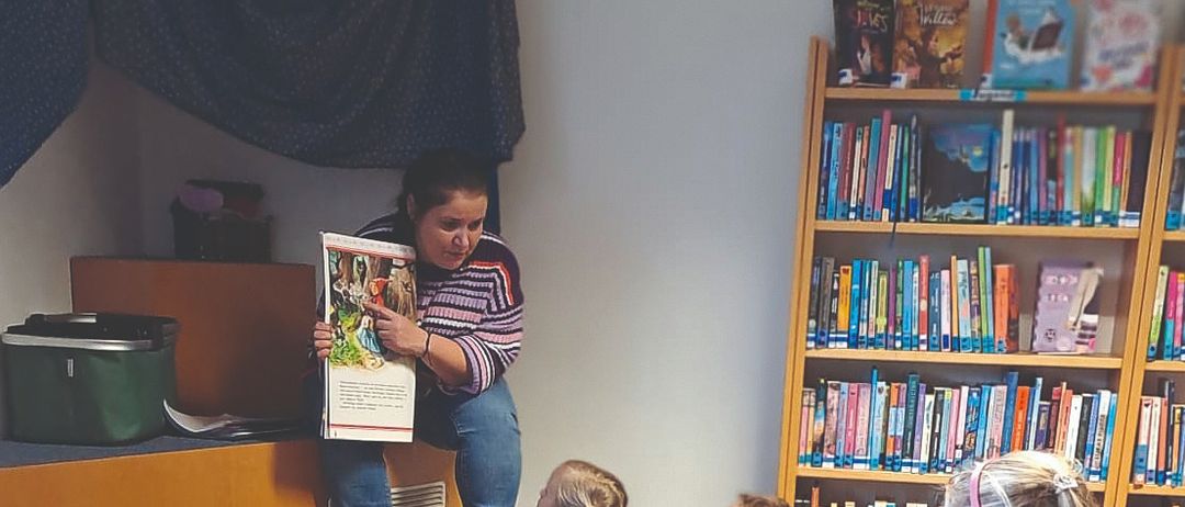 Eine Frau liest ein Buch vor einer Gruppe von Kindern, die in einem Kreis auf dem Boden in einer Bibliothek sitzen.