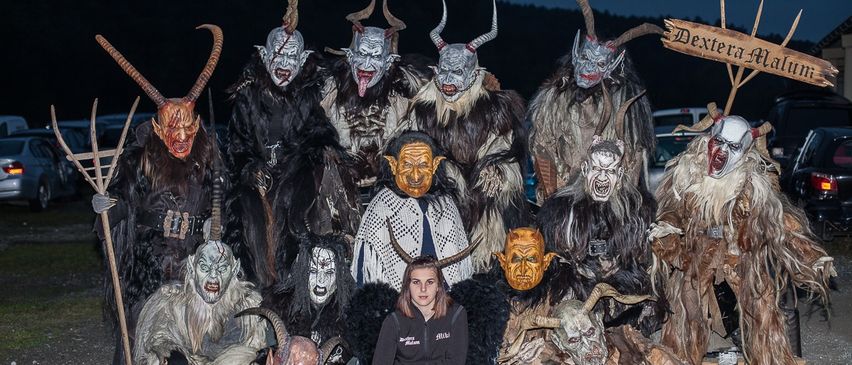 Eine Gruppe von Menschen in Kostümen posiert für ein Foto, einige mit Hörnern und Masken. Eine Frau in einer Jacke hockt in der Mitte. Der Ort scheint ein bewaldetes Gebiet zu sein.