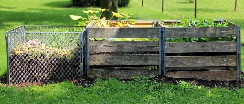 Zwei hölzerne Kompostbehälter sind in einem Hinterhof-Garten aufgestellt, einer mit Pflanzen und der andere möglicherweise leer. Ein Metallgitterzaun umgibt den Bereich.