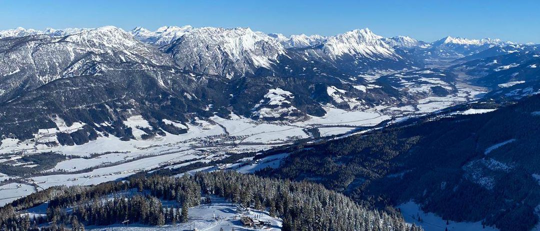 Luftaufnahme von verschneiten Bergen mit Kiefern, einem Tal und Skispuren im Vordergrund unter einem klaren blauen Himmel.