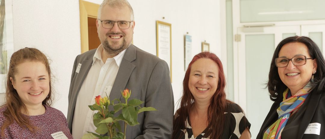 Vier Personen posieren für ein Foto in einem Raum mit weißen Wänden und einem weißen Tisch. Ein Mann steht hinter dem Tisch mit einem Blumenarrangement, und ein Schild auf dem Tisch sagt 'Willkommen bei uns!'.