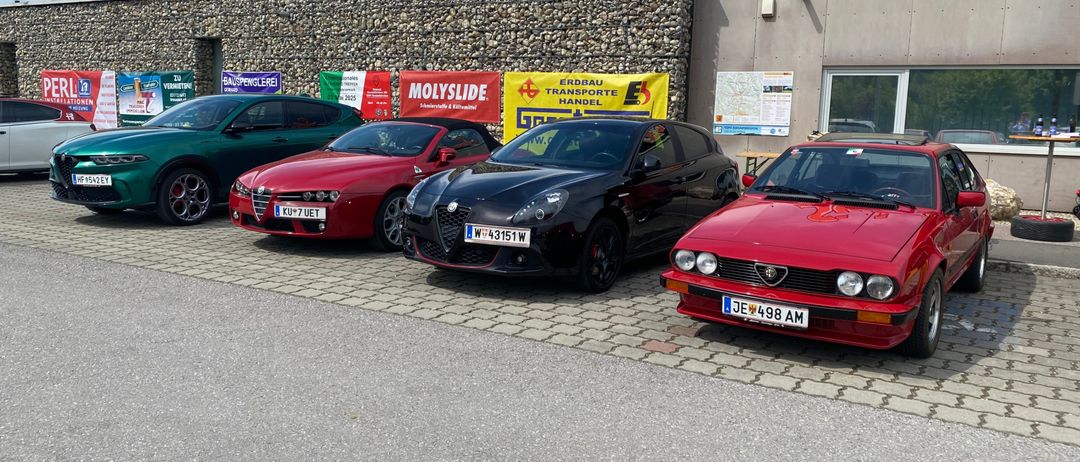 Mehrere Oldtimer sind vor einem Gebäude mit dem Schild 'Sinni' geparkt. Die Autos sind ein grüner, roter und schwarzer Alfa Romeo.