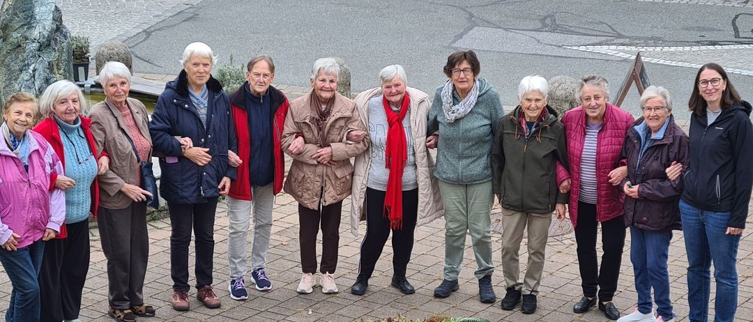 Eine Gruppe älterer Frauen steht lächelnd zusammen auf einem Ziegelweg, mit einer farbenfrohen Blumenanordnung in der Mitte. Sie tragen Jacken und Turnschuhe.