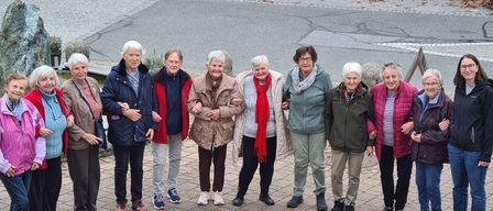 A group of women stands around a flower arrangement on a paved surface. A lantern is placed near the flowers.