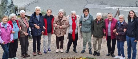 Eine Gruppe älterer Frauen steht vor einem Blumenkreis. Sie tragen Mäntel und Turnschuhe. In der Mitte des Kreises steht eine Laterne.