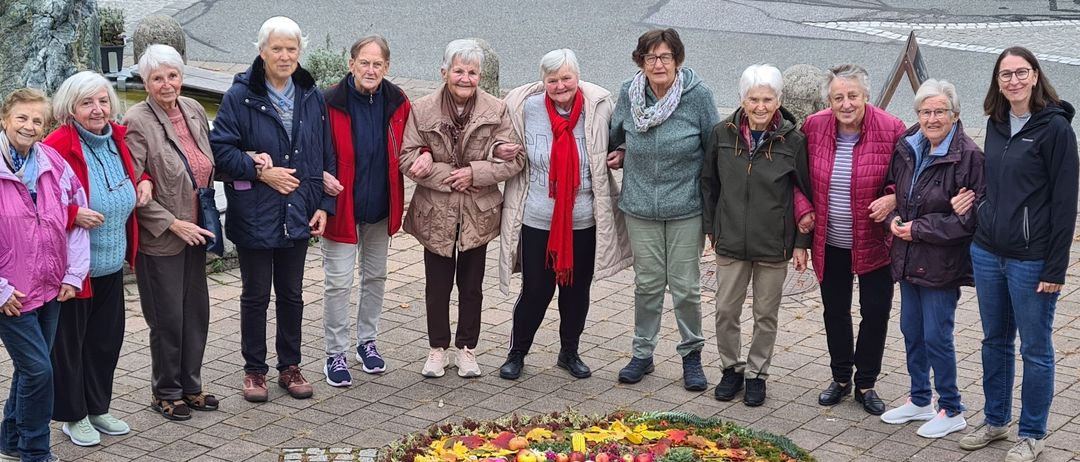Eine Gruppe älterer Frauen posiert um eine kreisförmige Blumenarrangement mit Herbstblättern und kleinen weißen Steinen. Sie stehen auf einem Ziegelweg in einem Außenbereich.