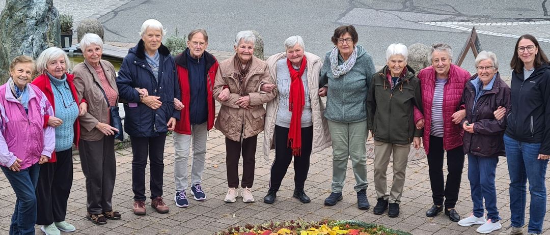 Eine Gruppe von Frauen in Herbstkleidung steht um einen bunten Kranz herum, möglicherweise für eine Gemeinschaftsveranstaltung oder Feier.