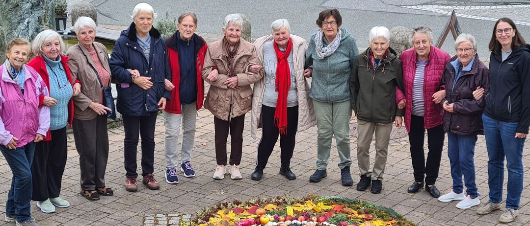 Eine Gruppe älterer Frauen steht um einen bunten Kranz aus Blumen und Früchten auf einer gepflasterten Oberfläche.