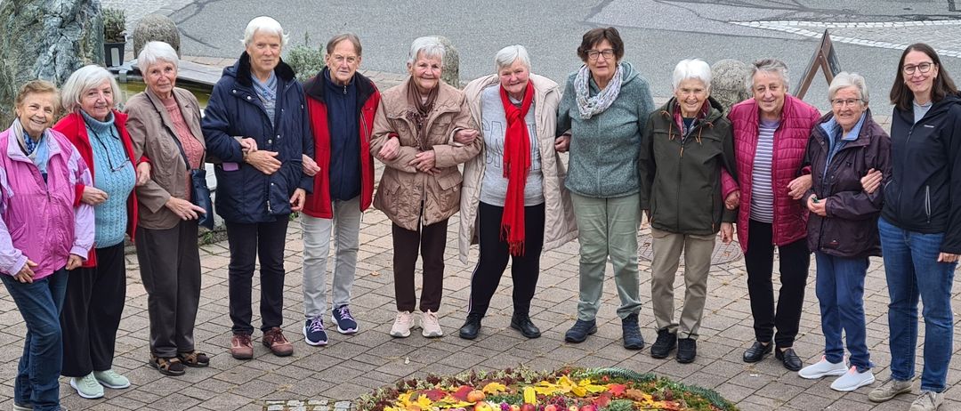 Eine Gruppe älterer Frauen steht um eine farbenfrohe Blumenarrangement mit Herbstblättern herum. Die Frauen sind warm in Mänteln und Stiefeln gekleidet. Ein Laternen steht auf der linken Seite.