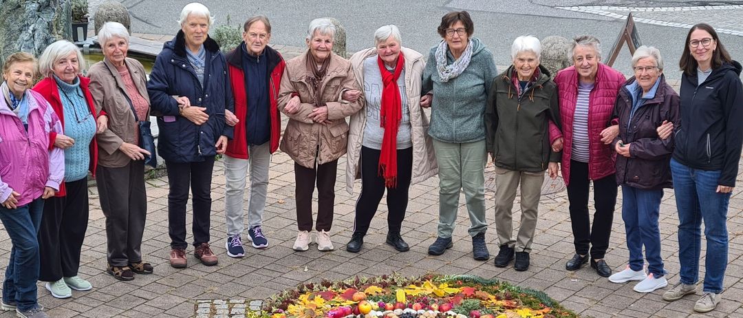 Eine Gruppe älterer Frauen steht um eine Blumendekoration im Freien. Sie sind warm gekleidet, was auf kühles Wetter hinweist.