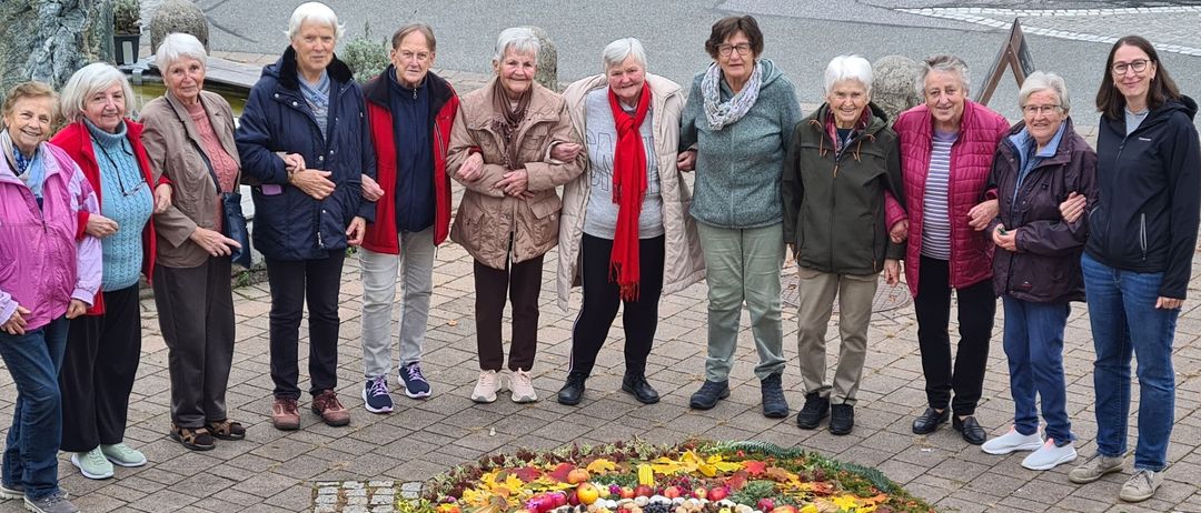 Eine Gruppe älterer Frauen posiert für ein Foto vor einer bunten Blumenanordnung auf einem gepflasterten Weg.