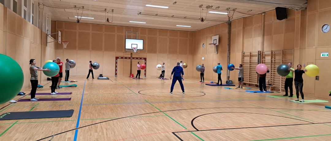 Sporthalle mit Basketballplatz, Personen in verschiedenen Positionen mit Trainingsbällen, einige in blauen Uniformen, und Yogamatten auf dem Boden verstreut.