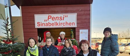 Eine Gruppe von Menschen steht vor einem kleinen Laden mit Weihnachtsdekoration, einschließlich eines Weihnachtsbaums und einem Schild, das 'Pensi's Sinabelkirchen' steht. Es gibt mehrere Erwachsene und ein Baby in einem Kinderwagen.