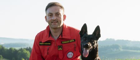 Ein Mann in einem roten Anzug kniet auf Gras mit einem schwarzen Hund, der einen roten Gurt trägt. Der Mann lächelt und der Hund hat die Zunge herausgestreckt.
