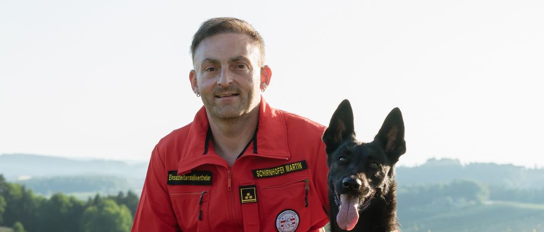 Ein Mann in roter Feuerwehruniform posiert mit einem schwarzen Hund auf einer Wiese mit einem Hintergrund aus Bergen.