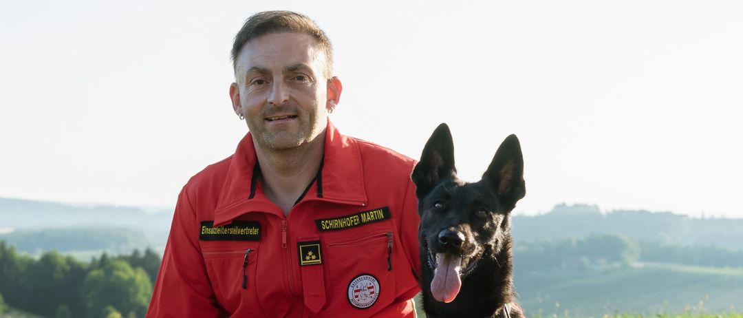 Ein Mann in einem roten Overall kniet mit einem schwarzen Hund auf dem Gras, beide tragen Sicherheitswesten. Hinter ihnen ein Hügel mit Bäumen und ein klarer Himmel.