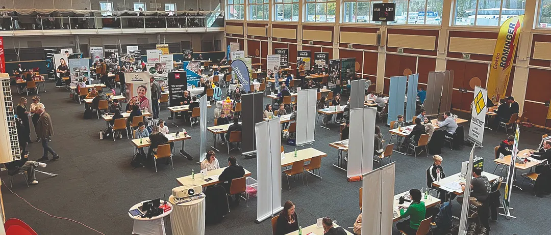 Ein Innenbereich mit vielen Tischen und Stühlen, gefüllt mit arbeitenden Menschen. Werbetafeln sind an der Wand angebracht und Lampen sind an der Decke montiert. Ein Projektor steht auf einem Tisch vorne.