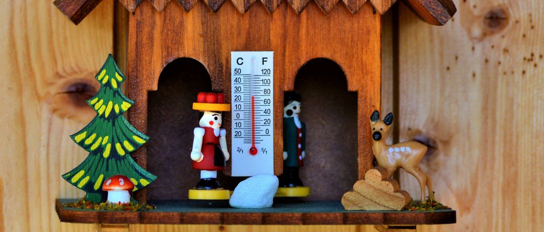 Ein holzfarbenes hausförmiges Objekt, möglicherweise eine Kuckucksuhr, zeigt ein Thermometer und Figuren von Menschen und Tieren im Inneren. Links stehen ein Weihnachtsbaum und ein Pilz.