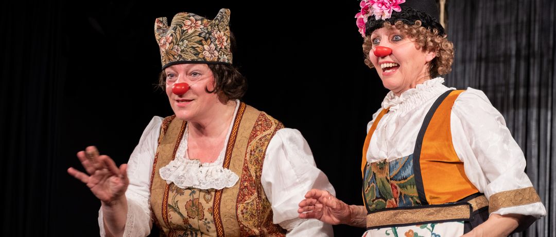 Two women dressed in colorful costumes, one with a floral crown, the other with a flowered hat and a red clown nose, smile and seem to be performing on a stage.