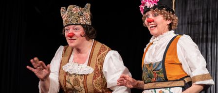 Two women dressed in colorful costumes, one with a floral crown, the other with a flowered hat and a red clown nose, smile and seem to be performing on a stage.