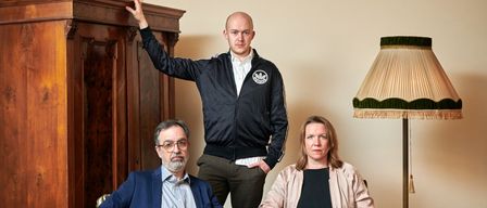 Three people pose in a room with a wooden cabinet, one leaning on it, and a lamp.