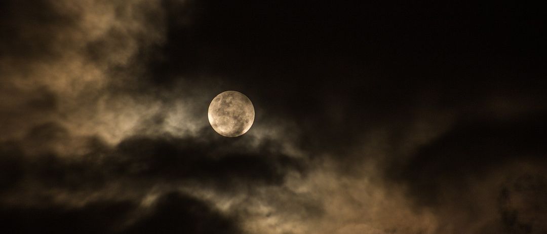 A full moon shines brightly through dark clouds on a night sky. The clouds appear thick and dense.