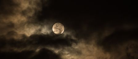 A full moon shines brightly through dark clouds on a night sky. The clouds appear thick and dense.