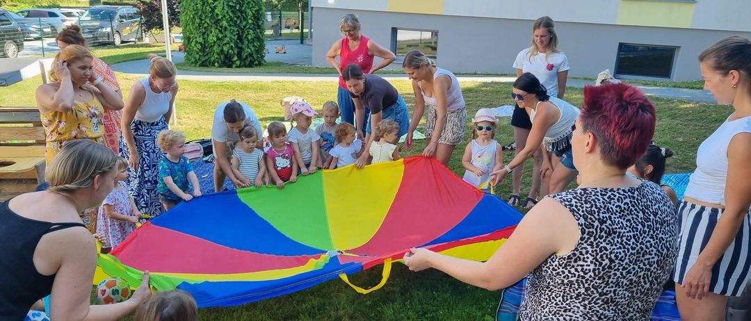 Mehrere Erwachsene und Kinder sind draußen. Sie halten einen großen, farbenfrohen Fallschirm. Eine Frau mit einem Leopardenmusterhemd hilft den Kindern. Sie befinden sich in der Nähe eines Gebäudes.