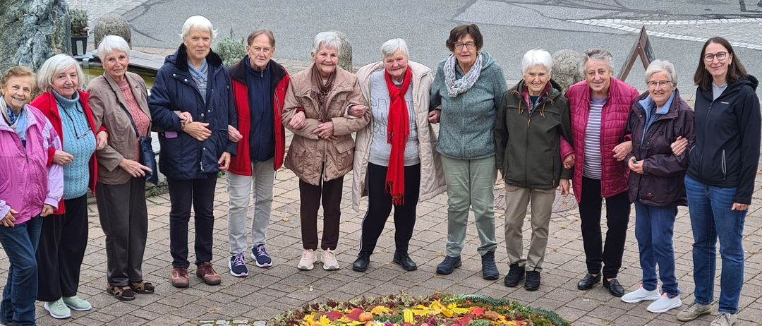 A group of women stand around a wreath of autumnal fruits and leaves. They are in a paved area.