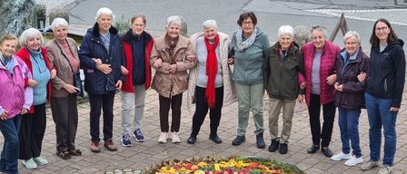 Eine Gruppe älterer Frauen steht um eine kreisförmige Blumenarrangement herum, wahrscheinlich für ein Foto, in einem Außenbereich.