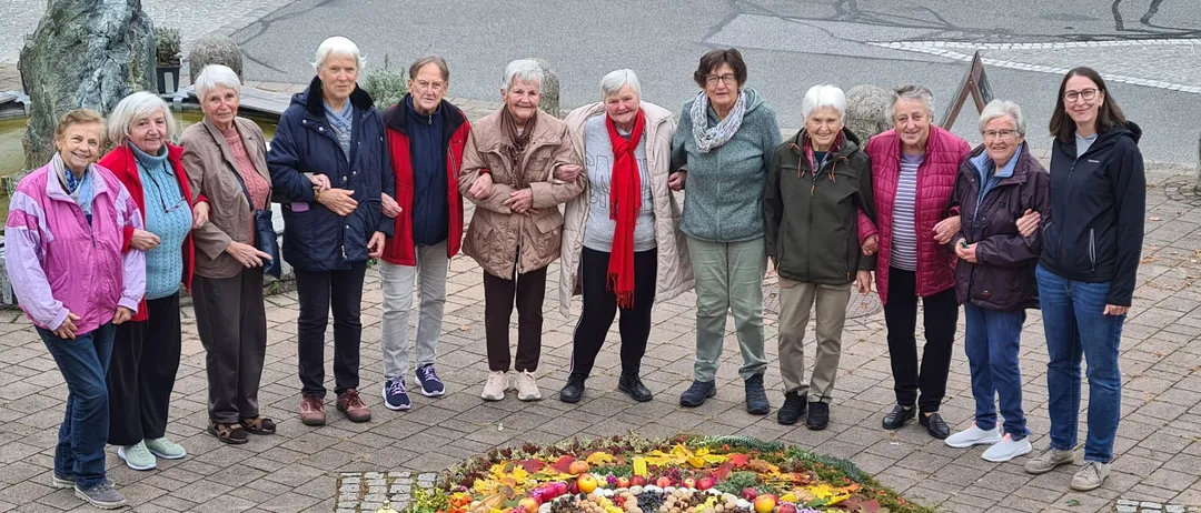 Eine Gruppe älterer Frauen steht um einen Kranz aus Herbstblättern, Früchten und Gemüse. Sie sind auf einem Ziegelboden.