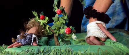Eine Frau in Kostüm, mit blauen Armschläuchen und Armbändern, ordnet rote Früchte auf einer grünen Oberfläche an. Eine Puppe mit einer blauen Armschlinge liegt auf dem Boden in der Nähe.