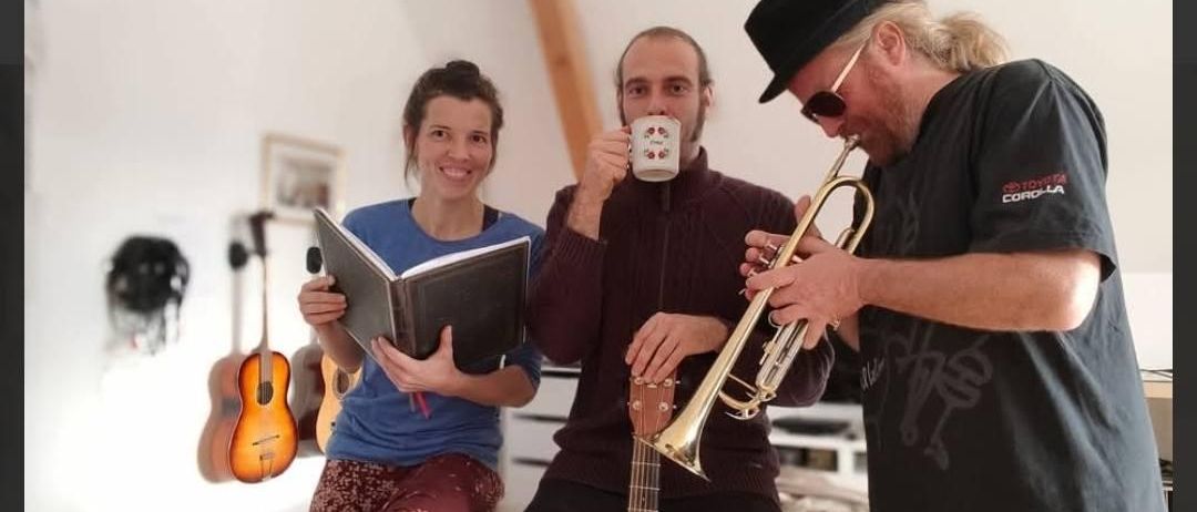 Drei Musiker posieren mit Instrumenten, einer trinkt, ein anderer spielt Trompete, und eine Frau hält ein Buch. Gitarren hängen an der Wand hinter ihnen.