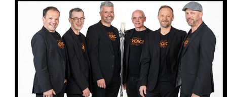 Six men in black suits stand together, smiling, with one man holding a microphone in front of them. They appear to be a musical group.