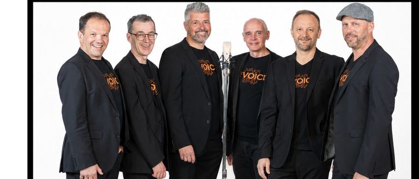 Sechs Männer in schwarzen Anzügen stehen zusammen und lächeln, einer von ihnen hält ein Mikrofon vor sich. Sie scheinen eine Musikgruppe zu sein.
