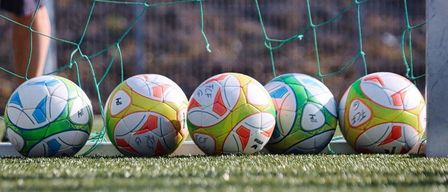 Fünf Fußballbälle sind vor einem grünen Tornetz auf einem Spielfeld aufgereiht.