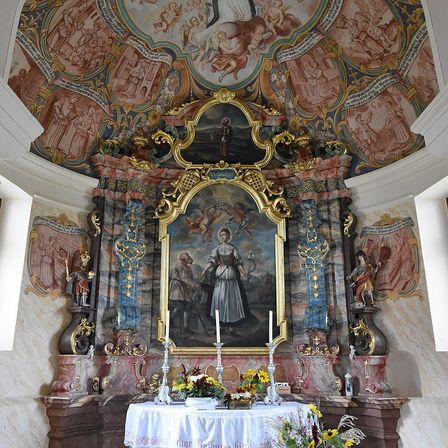 Ein Altar in der Kirche zeigt ein Gemälde einer Frau und eines Mannes, mit Kerzen und Blumen auf dem Altar und Fresken an den Wänden.