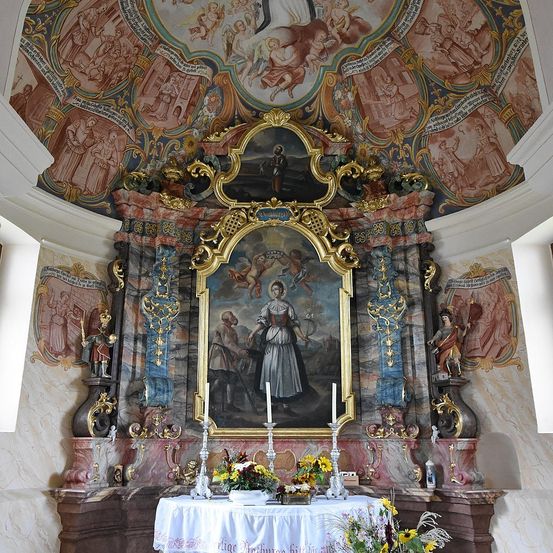 Ein Altar in der Kirche zeigt ein Gemälde einer Frau und eines Mannes, mit Kerzen und Blumen auf dem Altar und Fresken an den Wänden.