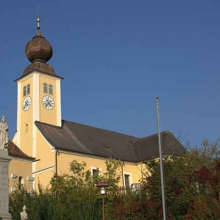 Eine gelbe Kirche mit einem grauen Dach, einem Glockenturm und zwei Uhren, die gegen einen klaren blauen Himmel steht.
